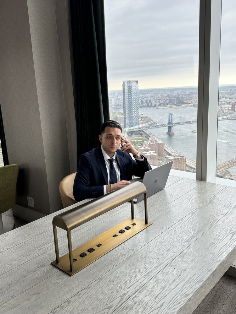 Migir Ilganayev at a desk on the phone, a laptop open in front of him and the East River behind.